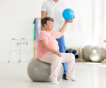 Older woman on an exercise ball holding another ball in her hand working on functional movement with a personal trainer