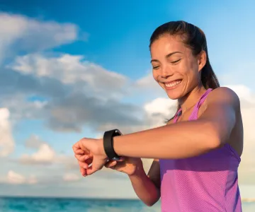 Woman in pink tank top looking at a fitness watch outside with clear waters and clouds in the background