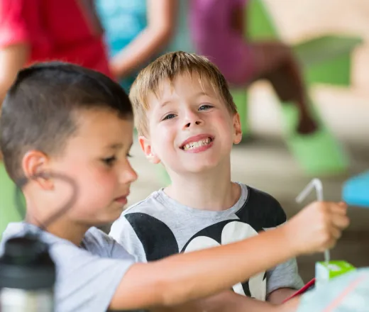 Cute boy looking at camera smiling while at lunch - friend is focused on his juice box