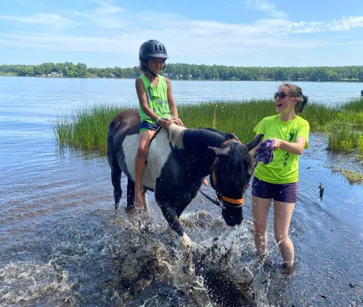 Camper wearing a helmet riding a horse in the water with staff guiding the horse for safety