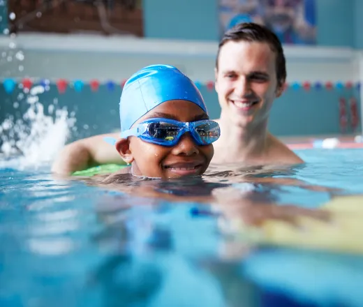 Male Swim Instructor Giving Boy Holding Float One To One Lesson In Pool